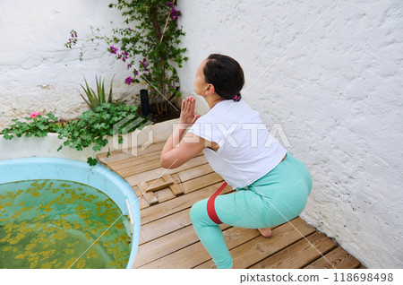 Woman practicing squat exercise with resistance band outdoors near a garden patio setting in bright athletic wear 118698498