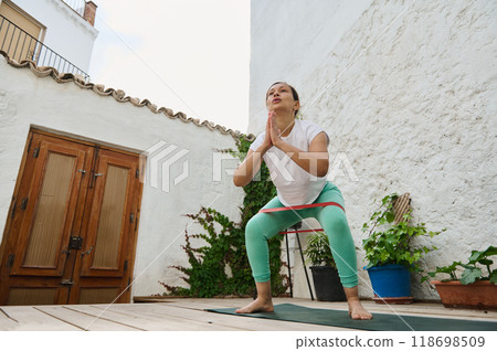 Woman practicing yoga in an outdoor courtyard, focusing on balance and flexibility in a serene environment with plants and rustic architecture Woman practicing yoga in an outdoor courtyard, focusing on balance and flexibility in a serene environment with plants and rustic architecture 118698509