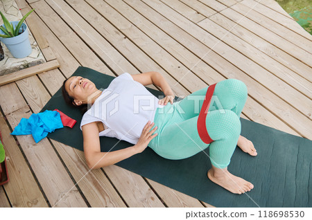 Woman lying on yoga mat outdoors using resistance band for exercise on a wooden deck in a peaceful setting Woman lying on yoga mat outdoors using resistance band for exercise on a wooden deck in a peaceful setting 118698530
