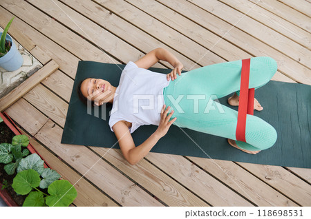 Woman exercising with resistance band on yoga mat outdoors, focused on strengthening and stretching muscles for improved fitness and wellness Woman exercising with resistance band on yoga mat outdoors, focused on strengthening and stretching muscles for improved fitness and wellness 118698531