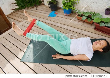 Woman practicing leg exercise with resistance band on yoga mat in outdoor garden setting for fitness and relaxation Woman practicing leg exercise with resistance band on yoga mat in outdoor garden setting for fitness and relaxation 118698542