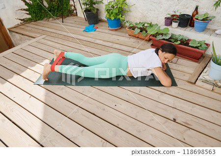 Woman exercising with resistance band on a wooden deck, focusing on fitness and relaxation in an outdoor setting, surrounded by potted plants and natural light 118698563
