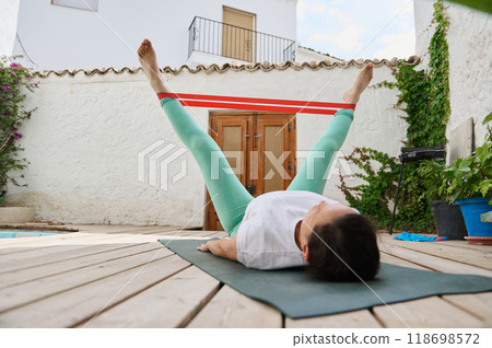 Woman exercising with resistance band outdoors performing leg workout on wooden deck emphasizing fitness and wellness routine. Woman exercising with resistance band outdoors performing leg workout on wooden deck emphasizing fitness and wellness routine. 118698572