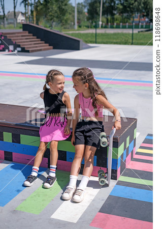 Two preschool-age girls relax and have fun chatting in a skatepark after skateboarding Two preschool-age girls relax and have fun chatting in a skatepark after skateboarding 118698648