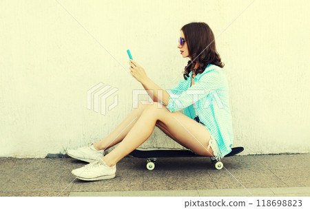 Modern young woman with smartphone sitting on skateboard on city street on white background Modern young woman with smartphone sitting on skateboard on city street on white background 118698823