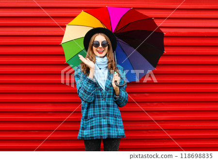 Fashion cheerful smiling woman holds colorful umbrella, black hat checkered jacket on red background 118698835