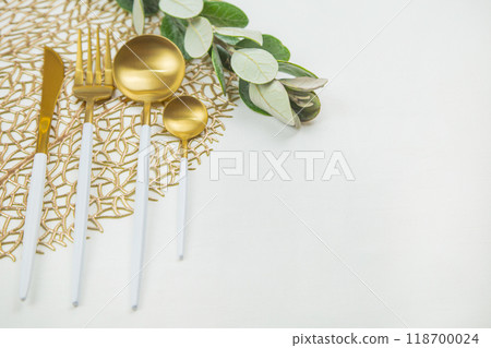 Cutlery: a knife, fork, spoon and a teaspoon of gold color with a white handle lie on a napkin of gold color, next to a green twig on a white background. The concept of food, culture and eating Cutlery: a knife, fork, spoon and a teaspoon of gold color with a white handle lie on a napkin of gold color, next to a green twig on a white background. The concept of food, culture and eating 118700024