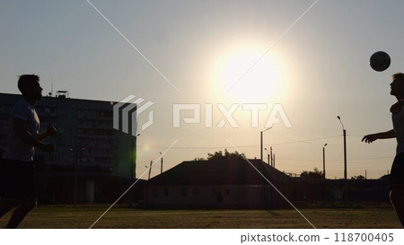 Silhouette of professional footballers kicking soccer ball with their heads on stadium at sunset. Two sportsmen showing tricks with ball while passing it to each other at field. Freestyle football 118700405