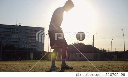 Professional footballer juggling soccer ball on stadium at sunset. Young man kicking ball at green field. Sportsman practicing tricks at meadow with sunlight at background. Freestyle football concept 118700406
