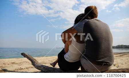 Romantic couple hugs during sitting on wooden log on the beach. Happy pair admiring a sea and landscape. Lovers spending time together at resort. Concept of vacation or holiday. Slow motion Romantic couple hugs during sitting on wooden log on the beach. Happy pair admiring a sea and landscape. Lovers spending time together at resort. Concept of vacation or holiday. Slow motion 118700425