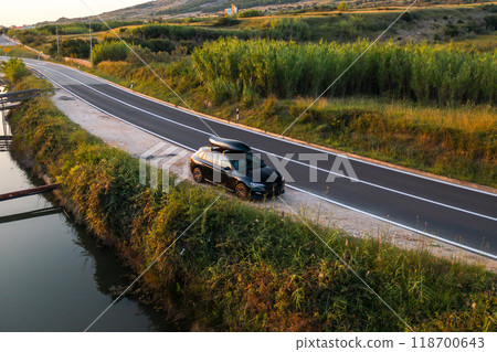 A car parked beside a tranquil river on a winding road during golden hour in the countryside. A car parked beside a tranquil river on a winding road during golden hour in the countryside. 118700643