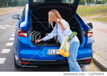 Young woman unloading groceries from the trunk of a blue electric car at a shopping center outdoor Young woman unloading groceries from the trunk of a blue electric car at a shopping center outdoor 118700645