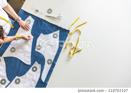 A woman dressmaker lays out pattern details on denim and cuts clothes. Top view. A woman dressmaker lays out pattern details on denim and cuts clothes. Top view. 118703107