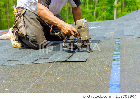 Roofer worker installing new bitumen shingles on roof used an air pneumatic nail gun. 118703904