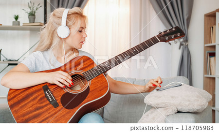 Guitar playing. Professional woman. Home lesson. Inspired lady in headphones tuning strings of musical instrument using mobile phone light room interior. 118705983