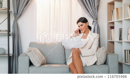Distance agreement. Female chief. Mobile chatting. Positive elegant woman talking smartphone holding paper documents sitting sofa light room interior. 118705986