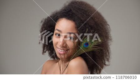 Health, beauty, tenderness and softness concept - beautiful African American woman with curly afro hairstyle and clean, healthy skin, smiles and touches her face with a bright colorful peacock feather Health, beauty, tenderness and softness concept - beautiful African American woman with curly afro hairstyle and clean, healthy skin, smiles and touches her face with a bright colorful peacock feather 118706010