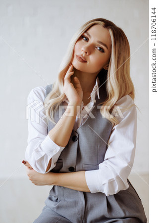 Young blonde woman in gray vest and trousers sits on black chair on white background.  118706144