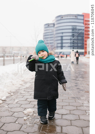 Boy child in a blue cap and a black coat is eating candy on a snowy sidewalk Boy child in a blue cap and a black coat is eating candy on a snowy sidewalk 118706845
