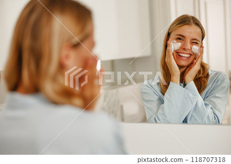 A woman is diligently applying her skincare products while looking into a mirror reflection A woman is diligently applying her skincare products while looking into a mirror reflection 118707318