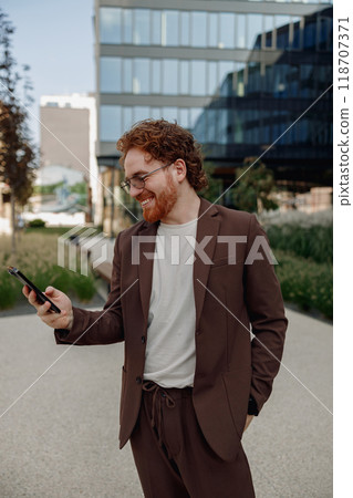 A stylish man in a brown suit uses his smartphone in an urban setting, embodying confidence 118707371