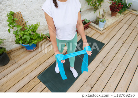 Woman practicing resistance band workout on wooden deck in outdoor garden setting Woman practicing resistance band workout on wooden deck in outdoor garden setting 118709250