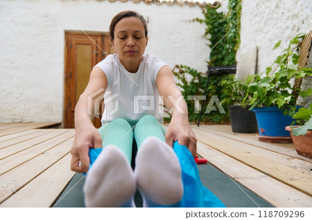 Woman practicing yoga in a peaceful outdoor setting, focusing on stretching exercises for relaxation and flexibility on a wooden deck surrounded by plants. Woman practicing yoga in a peaceful outdoor setting, focusing on stretching exercises for relaxation and flexibility on a wooden deck surrounded by plants. 118709296