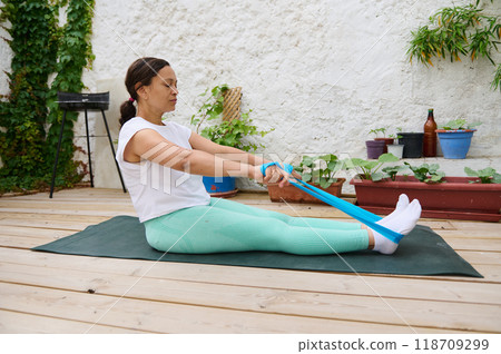 Woman practicing outdoor yoga with resistance band on wooden deck surrounded by potted plants for fitness and wellness 118709299