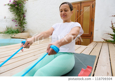 Woman practicing yoga with resistance bands on a mat in a peaceful outdoor setting, focusing on balance and mindfulness to enhance her wellness routine. Woman practicing yoga with resistance bands on a mat in a peaceful outdoor setting, focusing on balance and mindfulness to enhance her wellness routine. 118709304