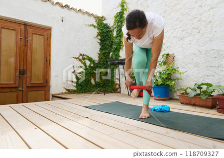 Woman practicing yoga with resistance band on a wooden deck surrounded by greenery Woman practicing yoga with resistance band on a wooden deck surrounded by greenery 118709327