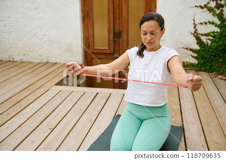 Woman performing resistance band exercises on wooden deck for home fitness routine Woman performing resistance band exercises on wooden deck for home fitness routine 118709635