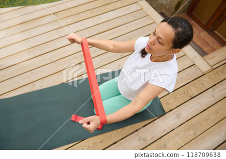 Woman practicing resistance band exercises on a wooden deck for fitness and strength training outdoors Woman practicing resistance band exercises on a wooden deck for fitness and strength training outdoors 118709638