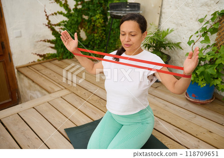Woman practicing resistance band exercise outdoors on wooden deck surrounded by greenery 118709651