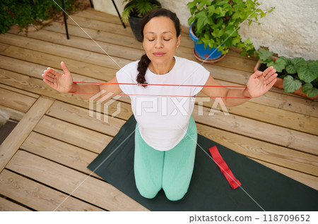 Woman practicing yoga with resistance band on a wooden deck while meditating. Indoor fitness and mindfulness for mental health and physical wellbeing. Woman practicing yoga with resistance band on a wooden deck while meditating. Indoor fitness and mindfulness for mental health and physical wellbeing. 118709652