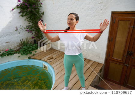 Woman exercising with resistance band on wooden deck by small pool in a serene garden setting 118709657