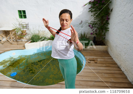 Woman practicing resistance band exercise outdoors by a pool with lush garden background on a sunny day 118709658