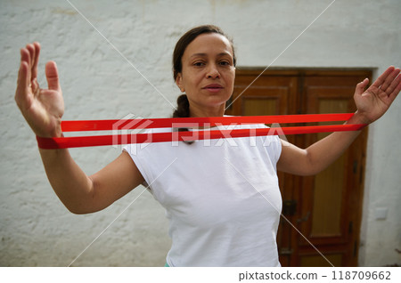 Confident woman practicing resistance band exercises outdoors for fitness and strength training in a relaxed setting Confident woman practicing resistance band exercises outdoors for fitness and strength training in a relaxed setting 118709662