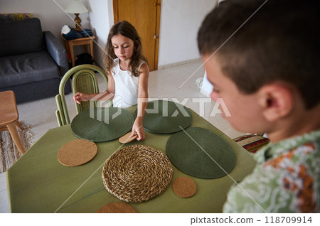 Young girl and boy set a dining table with green placemats and cork coasters in a cozy home environment. Young girl and boy set a dining table with green placemats and cork coasters in a cozy home environment. 118709914