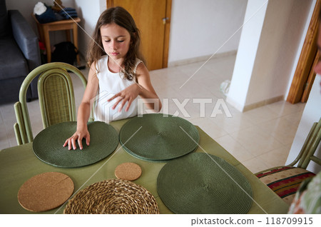 Young girl setting the dining table with round green placemats in a cozy home interior setting Young girl setting the dining table with round green placemats in a cozy home interior setting 118709915
