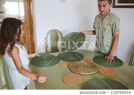 Children preparing dining table with green placemats in a cozy dining room setting Children preparing dining table with green placemats in a cozy dining room setting 118709916