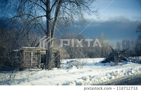 Old wooden hut in snowy landscape, logs in background, under dramatic sky. Cold air creates peaceful winter scene. Old abandoned hut against snowy field. Old wooden hut in snowy landscape, logs in background, under dramatic sky. Cold air creates peaceful winter scene. Old abandoned hut against snowy field. 118712718