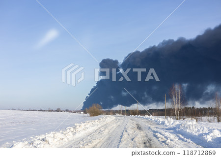 A large fire accompanied by thick smoke. A large black column of smoke rises above the horizon on a clear winter day, creating a sharp contrast against the blue sky and snow-covered fields. 118712869
