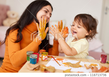 happy little child girl and mother spending good time painting hands and autumn yellow leaves together. smiling mom and baby showing hands in yellow paint happy little child girl and mother spending good time painting hands and autumn yellow leaves together. smiling mom and baby showing hands in yellow paint 118713736