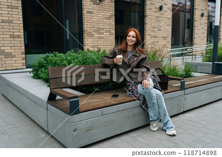 Portrait of joyful red-haired woman enjoying peaceful coffee break sitting on city bench in autumn day. Calm redhead female escaping urban rush for casual moment, enjoying free time in European city. Portrait of joyful red-haired woman enjoying peaceful coffee break sitting on city bench in autumn day. Calm redhead female escaping urban rush for casual moment, enjoying free time in European city. 118714898