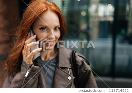 Closeup of beautiful young woman with long red hair and freckles chatting on smartphone outdoors, smiling looking at camera. Cute redhead female dressed in leather jacket enjoying conversation outside Closeup of beautiful young woman with long red hair and freckles chatting on smartphone outdoors, smiling looking at camera. Cute redhead female dressed in leather jacket enjoying conversation outside 118714931