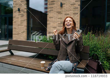 Cheerful redhead woman receiving unexpected good news in social media app using smartphone sitting on urban bench on summer day. Happy female celebrating success with joy and positive vibes. 118714933