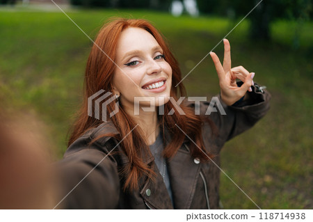 Portrait of positive young woman with long red hair smiling happily looking at camera, taking selfie in park, showing peace sign. Happy redhead female shooting selfie flashing peace sign. 118714938