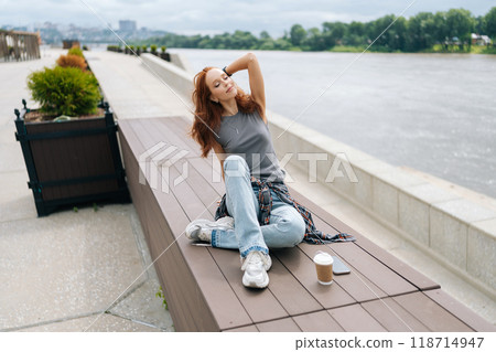 High-angle view of serene pretty young woman sitting on wooden platform by river, enjoying peaceful moment with coffee, cloudy sky and calm water creating serene atmosphere. High-angle view of serene pretty young woman sitting on wooden platform by river, enjoying peaceful moment with coffee, cloudy sky and calm water creating serene atmosphere. 118714947
