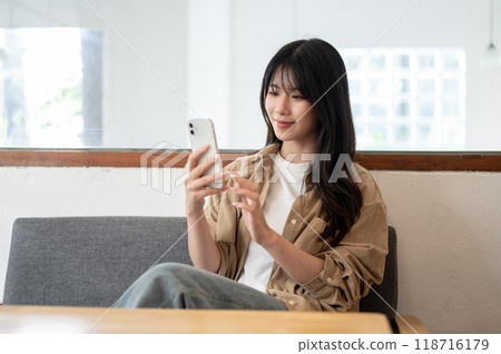 A beautiful, positive Asian woman is sitting in a coffee shop or living room, using her smartphone. 118716179