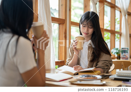 Two cheerful Asian college students are enjoying coffee while reading together in a coffee shop. 118716193
